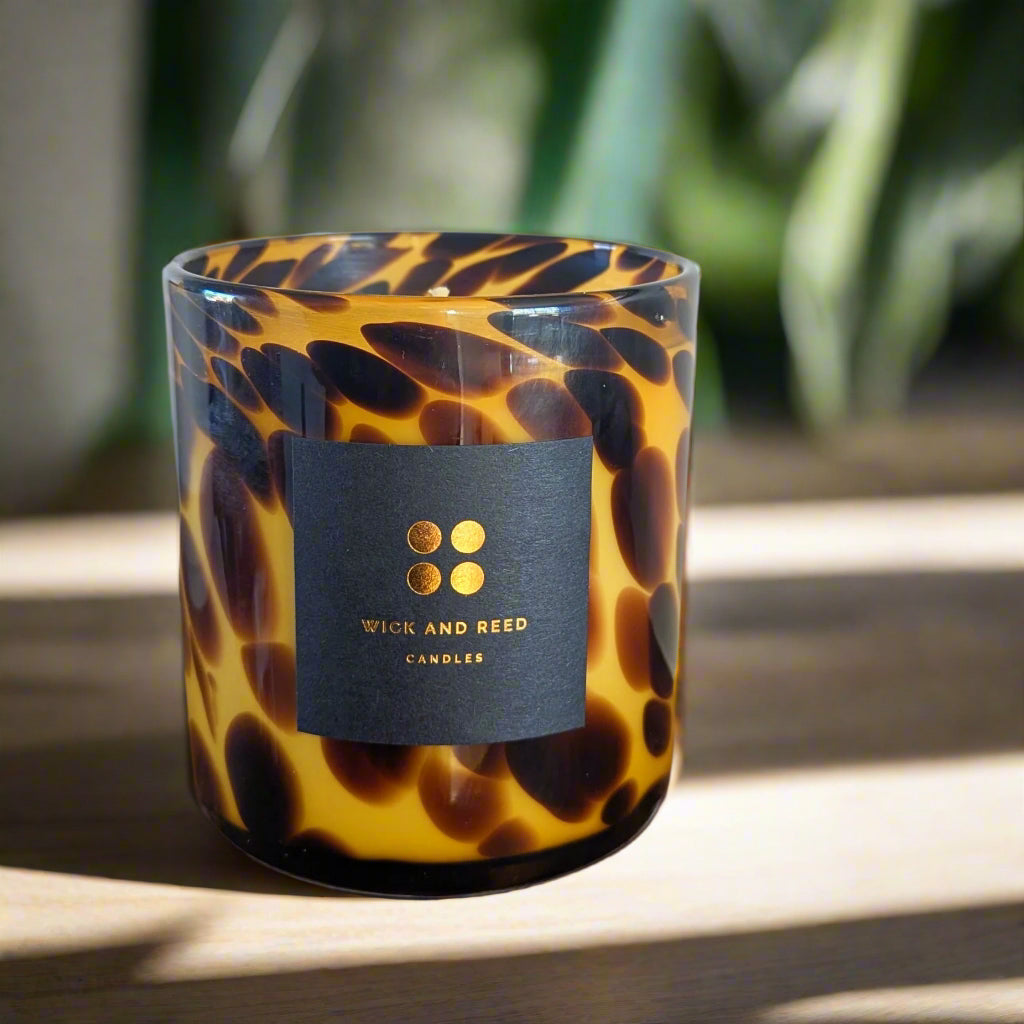 A candle with a black label that reads 'WICK AND REED CANDLES' in a patterned glass holder.